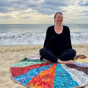 Tannia Joy sitting on her Sacred Space Hand Quilted Wheel on the beach in Mexico. She has so much joy and a big smile on her face as waves crash behind her.